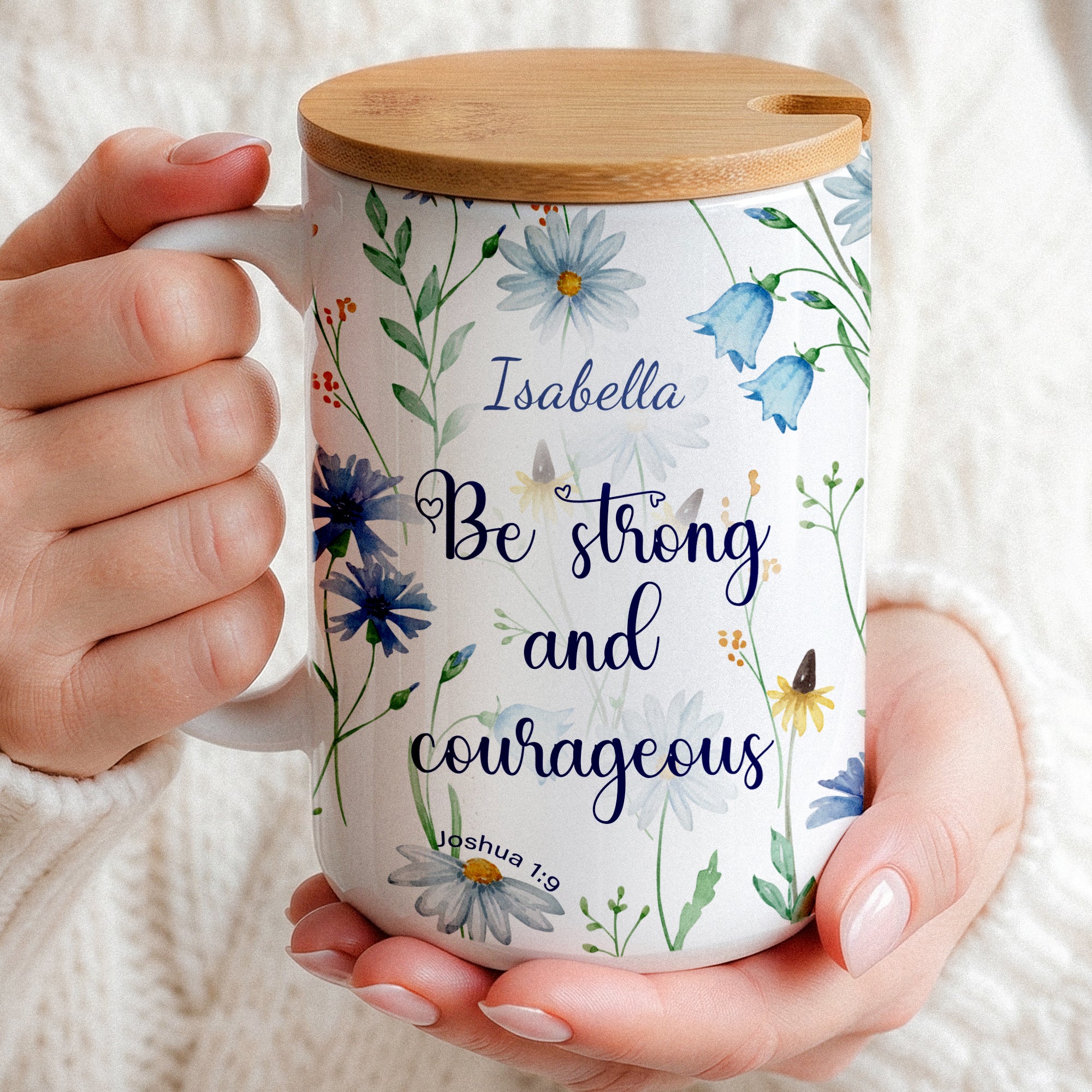 Personalized white ceramic mug featuring a blue watercolor wildflower design and the Christian bible verse Joshua 1:9 "Be strong and courageous" with a custom name. Available in an 11oz classic coffee cup style or an elegant 15oz premium gift set with a wooden bamboo lid and a golden flower spoon on a bright, minimalist marble background.