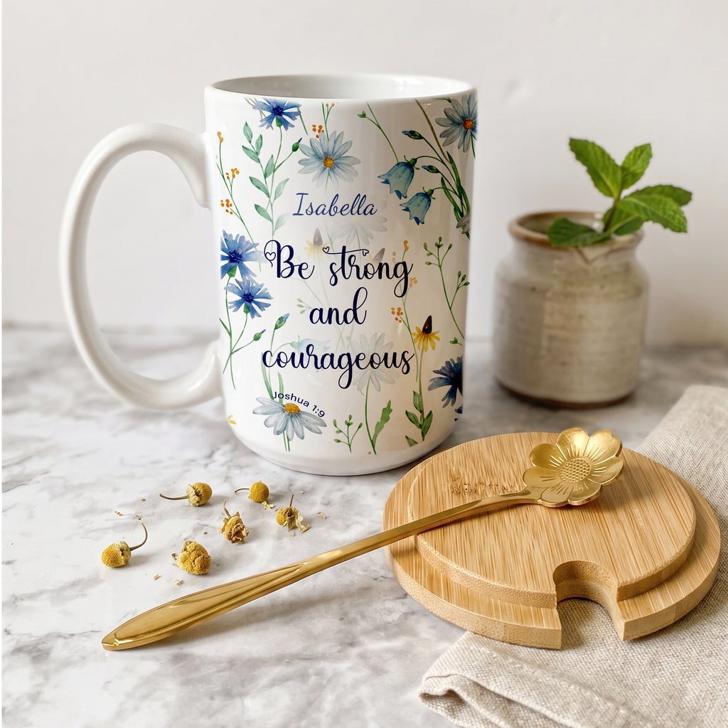 Personalized white ceramic mug featuring a blue watercolor wildflower design and the Christian bible verse Joshua 1:9 "Be strong and courageous" with a custom name. Available in an 11oz classic coffee cup style or an elegant 15oz premium gift set with a wooden bamboo lid and a golden flower spoon on a bright, minimalist marble background.