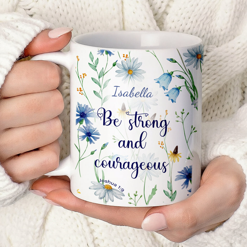 Personalized white ceramic mug featuring a blue watercolor wildflower design and the Christian bible verse Joshua 1:9 "Be strong and courageous" with a custom name. Available in an 11oz classic coffee cup style or an elegant 15oz premium gift set with a wooden bamboo lid and a golden flower spoon on a bright, minimalist marble background.