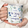 Personalized white ceramic mug featuring a blue watercolor wildflower design and the Christian bible verse Joshua 1:9 "Be strong and courageous" with a custom name. Available in an 11oz classic coffee cup style or an elegant 15oz premium gift set with a wooden bamboo lid and a golden flower spoon on a bright, minimalist marble background.