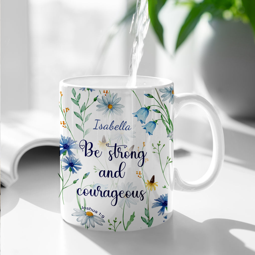Personalized white ceramic mug featuring a blue watercolor wildflower design and the Christian bible verse Joshua 1:9 "Be strong and courageous" with a custom name. Available in an 11oz classic coffee cup style or an elegant 15oz premium gift set with a wooden bamboo lid and a golden flower spoon on a bright, minimalist marble background.
